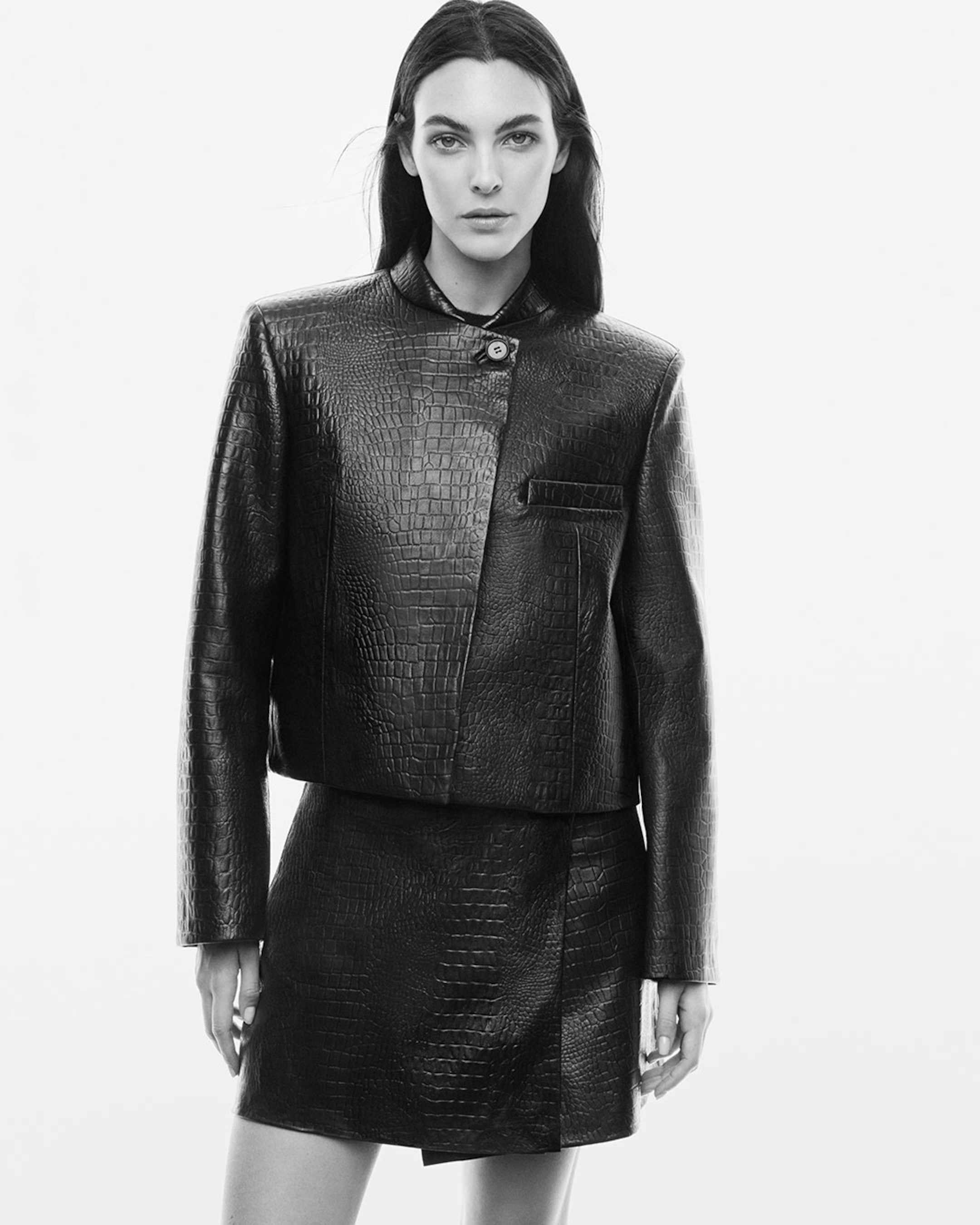 A woman in a textured black leather jacket and matching skirt, standing in a confident pose against a plain white background.