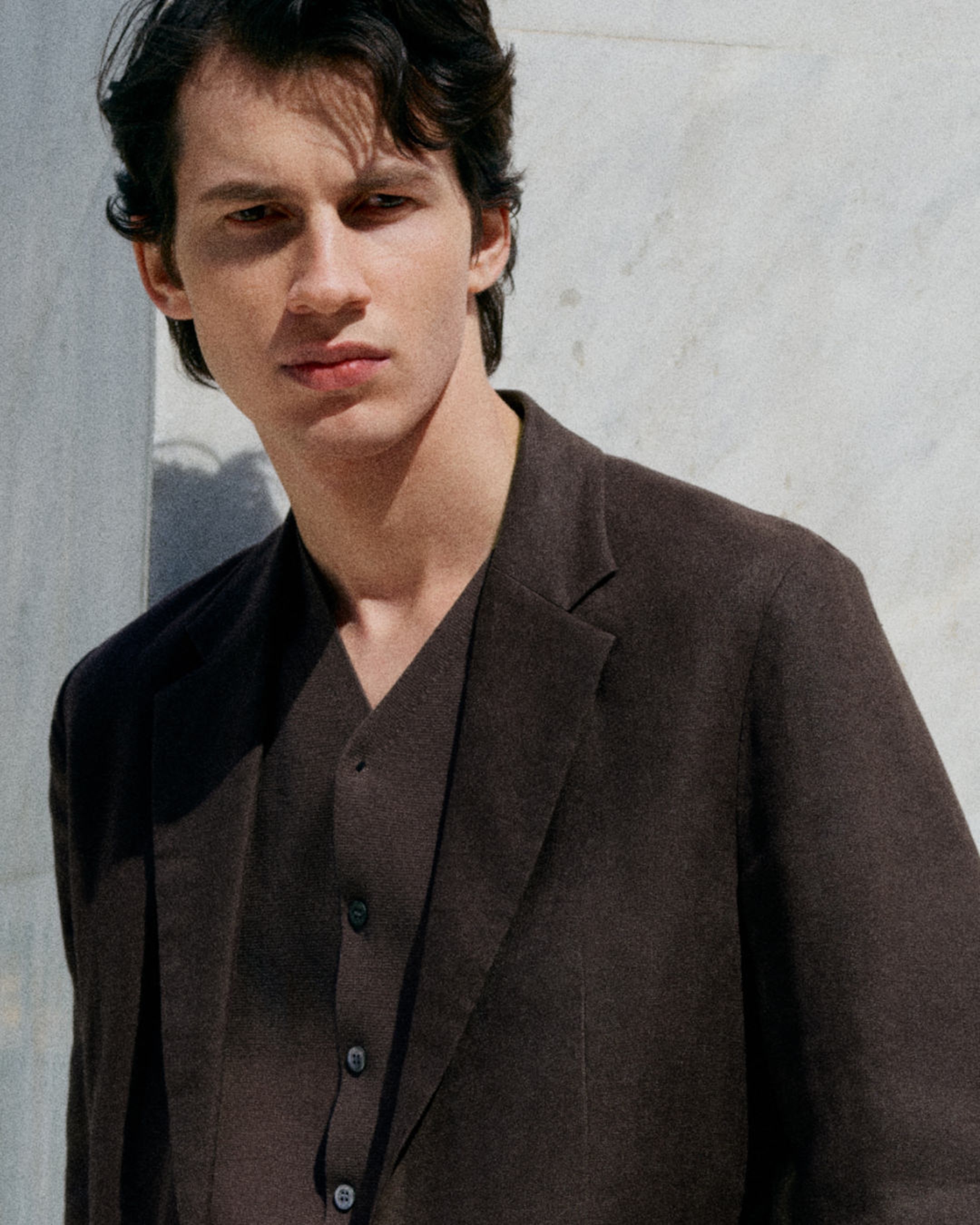 A person in a dark suit stands against a light-colored wall, looking slightly to the side, with short dark hair and a neutral expression.