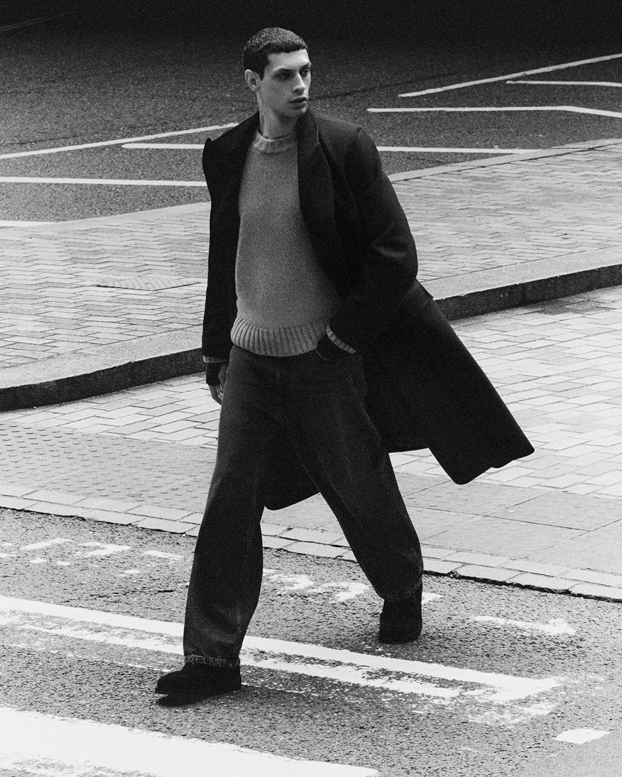 A person in a winter coat and sweater walks confidently across a street, with a shadowed background.