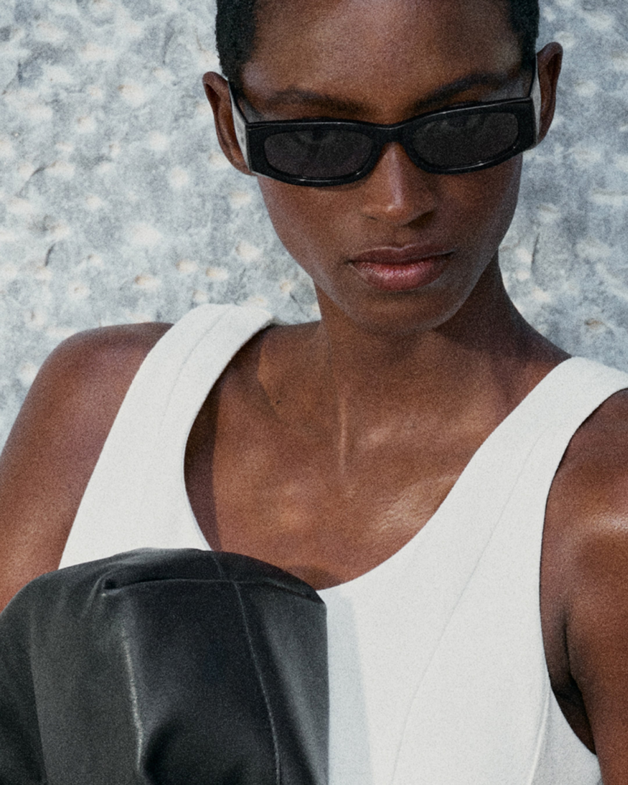 Person wearing black sunglasses, white sleeveless top, and holding a black item, against a textured light background.