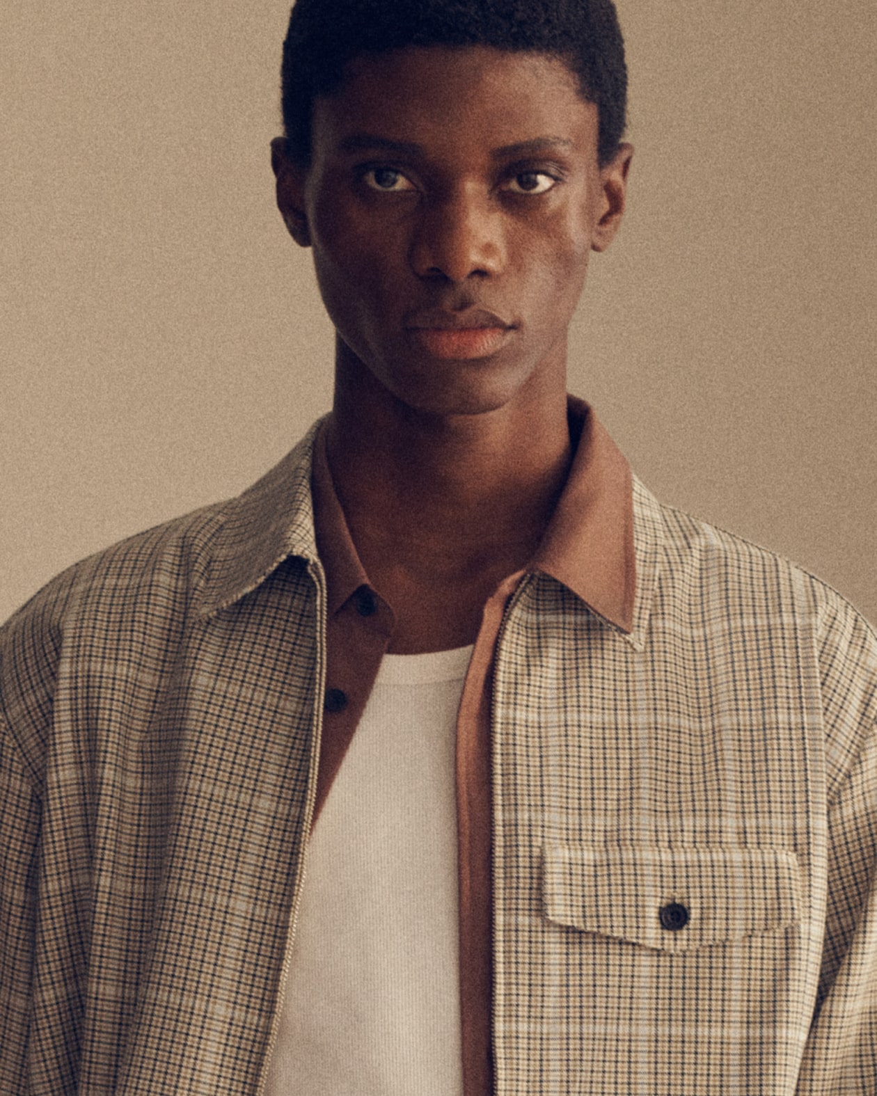A person wearing a plaid shirt over a white T-shirt, looking directly at the camera against a neutral background.