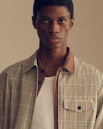 Placeholder for: A person wearing a plaid shirt over a white T-shirt, looking directly at the camera against a neutral background.