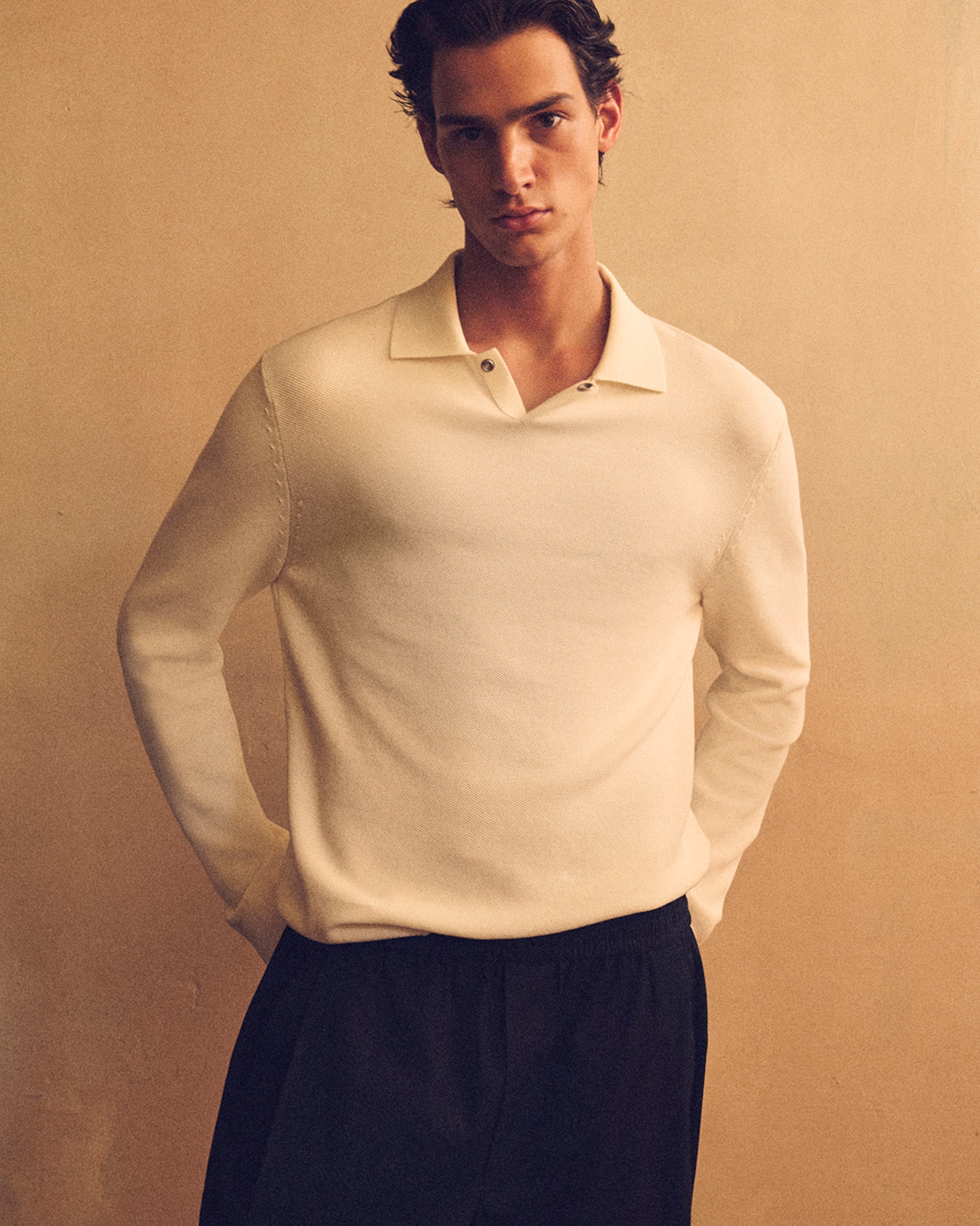 A person stands against a neutral background, wearing a cream-colored long-sleeve shirt and dark pants, with hands in their pockets.