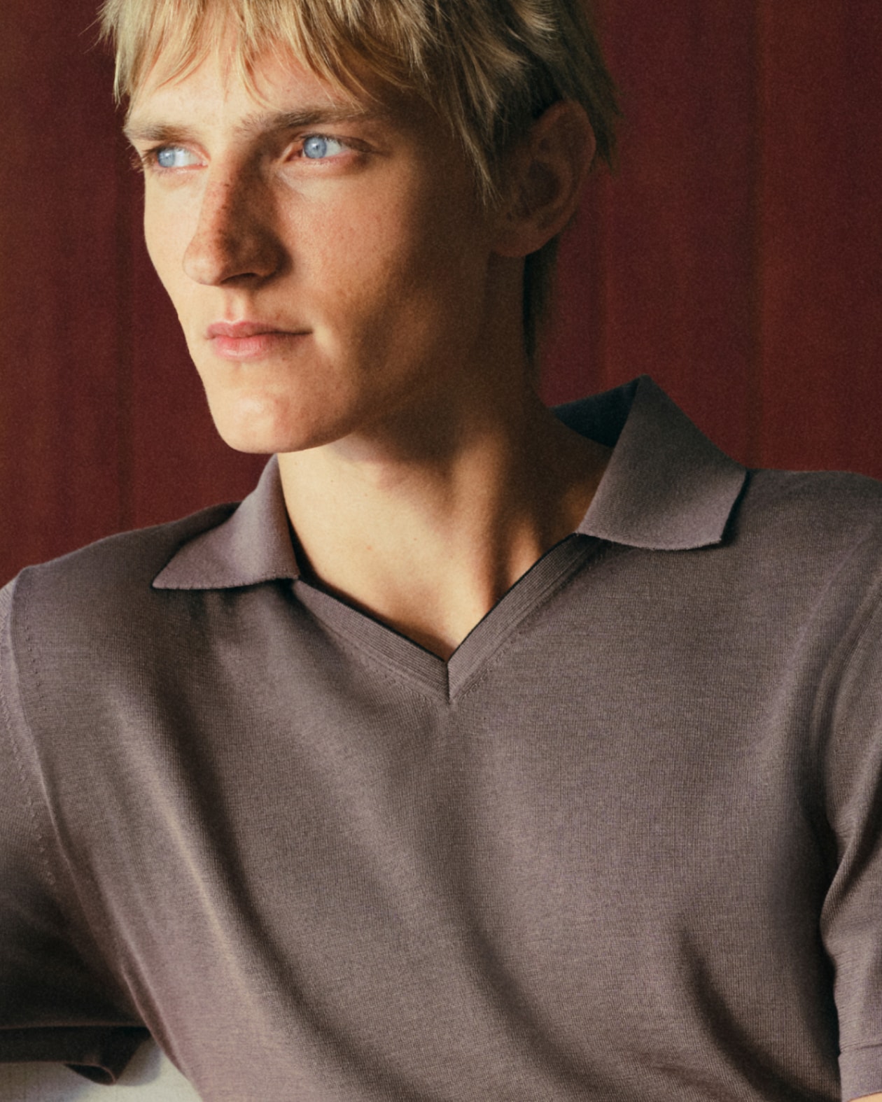 A person with blond hair wearing a brown shirt looks to the side with a thoughtful expression, set against a dark background.