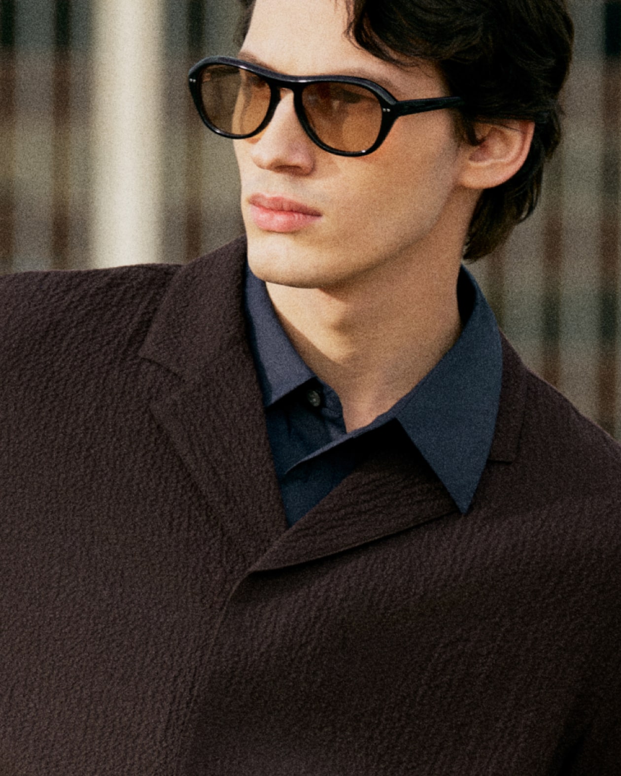 A person wearing large sunglasses and a textured dark coat over a blue shirt, looking to the side with a neutral expression.
