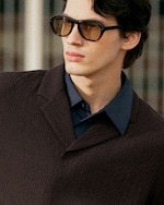 Placeholder for: A person wearing large sunglasses and a textured dark coat over a blue shirt, looking to the side with a neutral expression.
