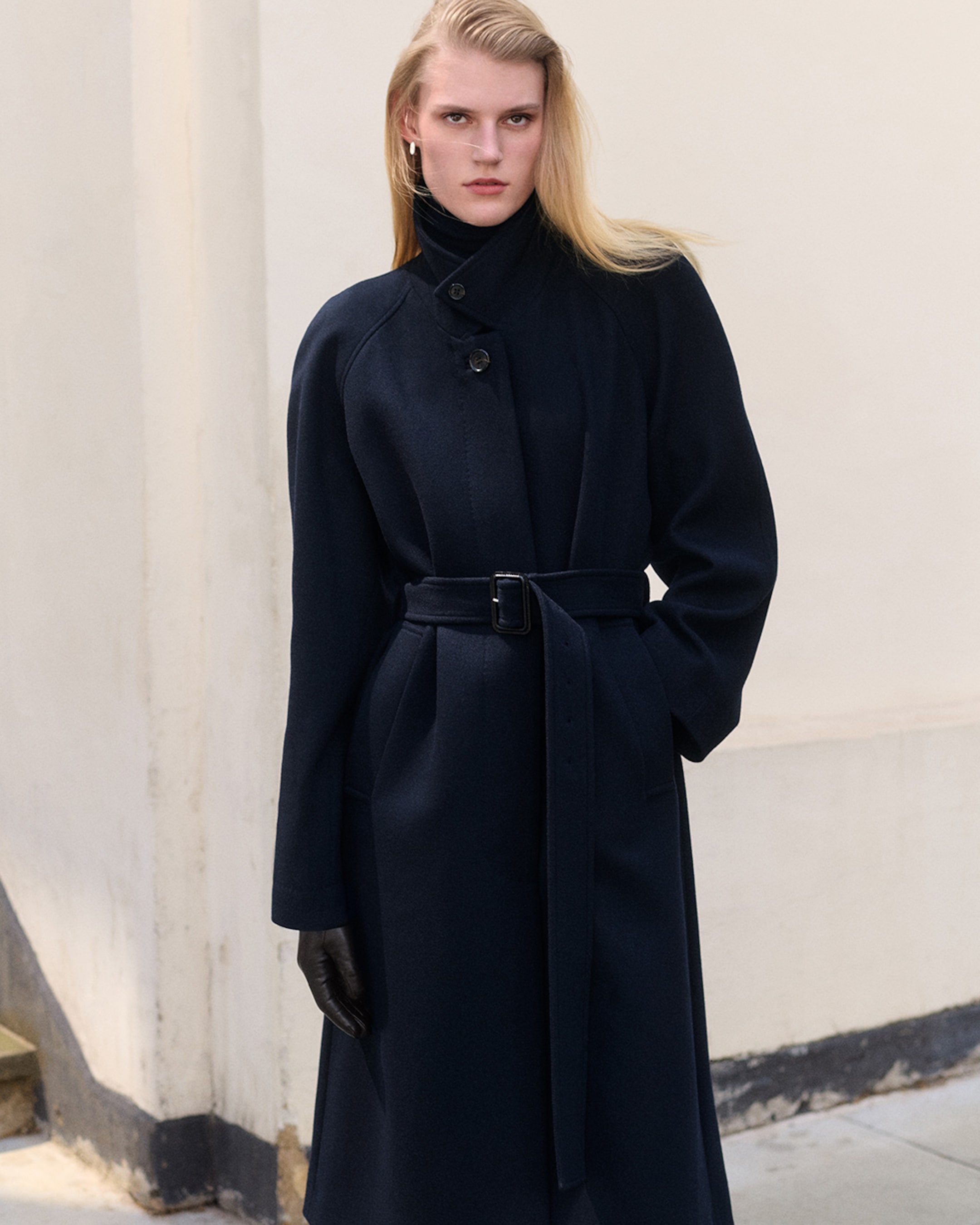 A person wearing a dark belted coat and gloves stands confidently in front of a light-colored wall.