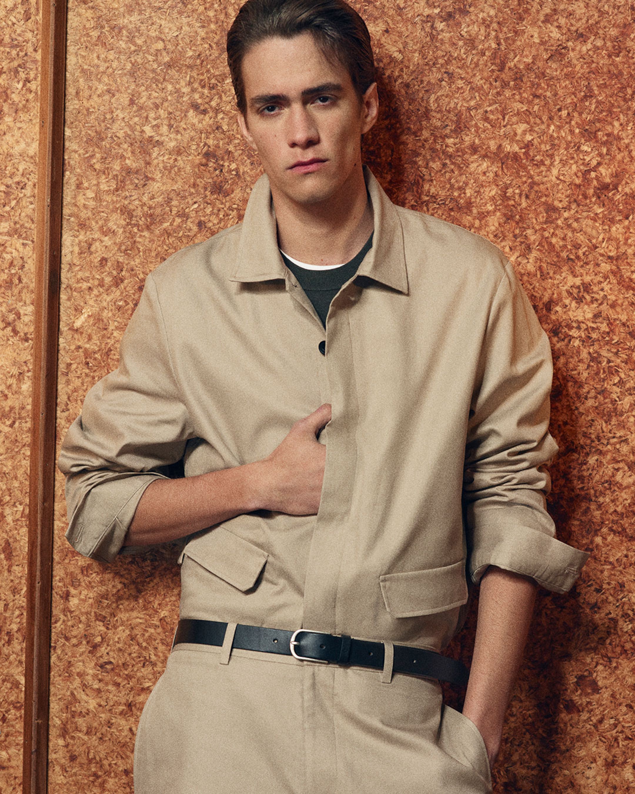 A person wearing a beige jacket with black belt against a textured brown background, looking directly at the camera with a neutral expression.
