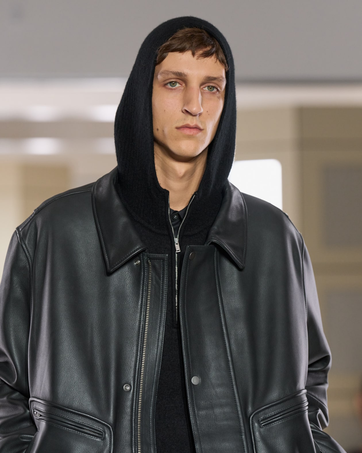 A person wearing a black hooded jacket with a leather exterior, standing indoors.