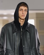 Placeholder for: A person wearing a black hooded jacket with a leather exterior, standing indoors.
