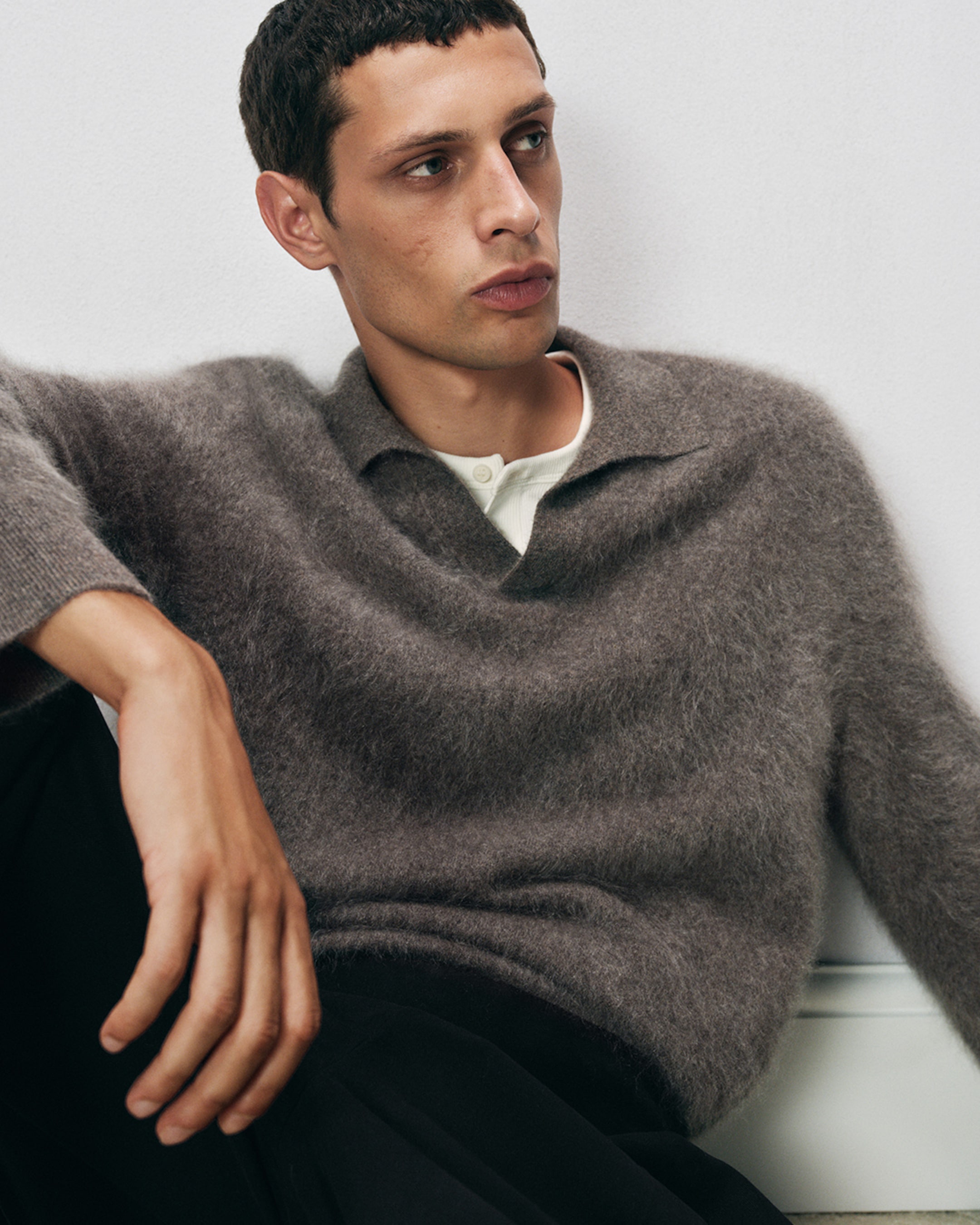Man wearing a fuzzy brown sweater over a cream shirt, seated, gazing to the side against a plain background.