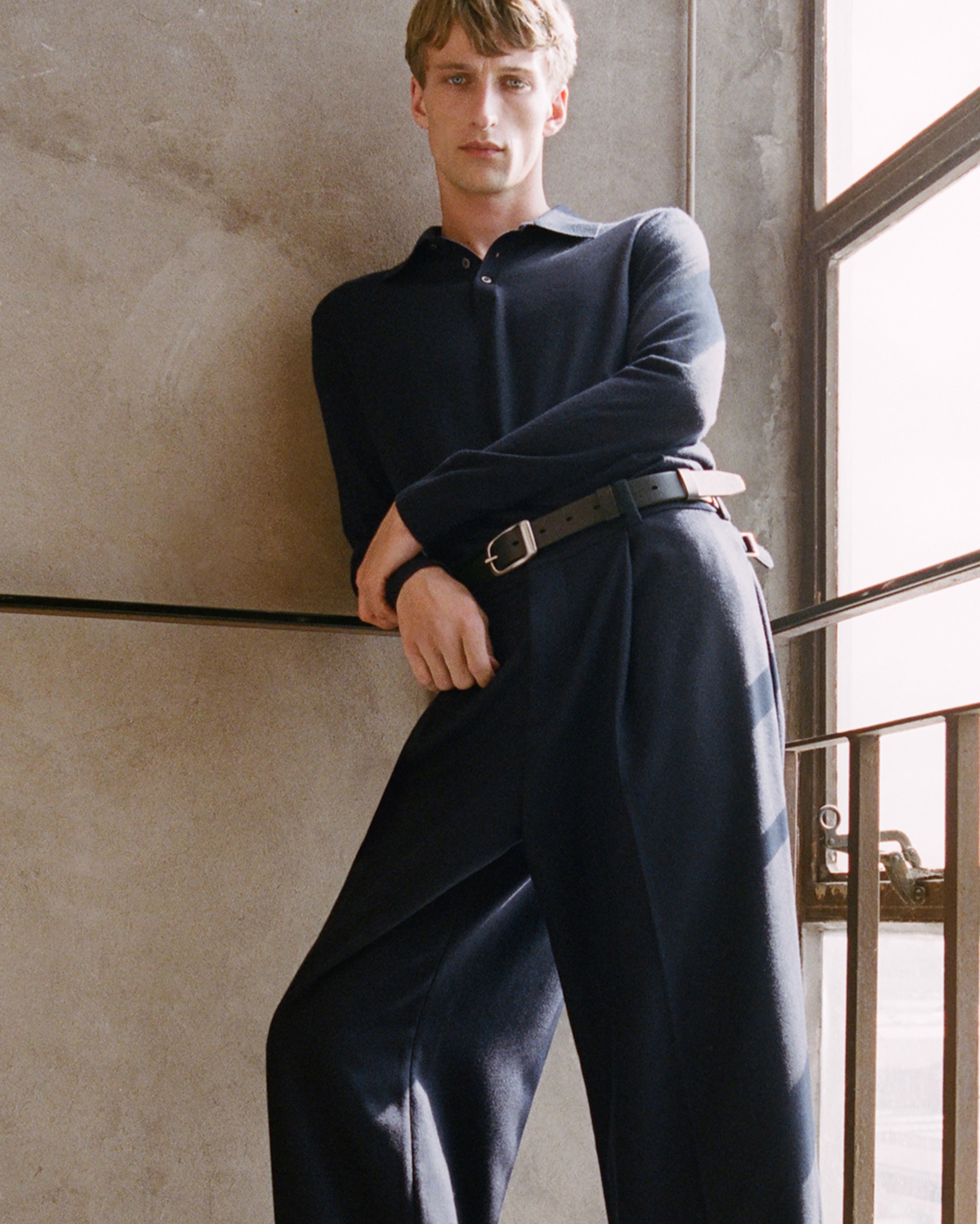 Person in a navy outfit leans against a textured wall by a window with metal bars, gazing forward with a neutral expression.