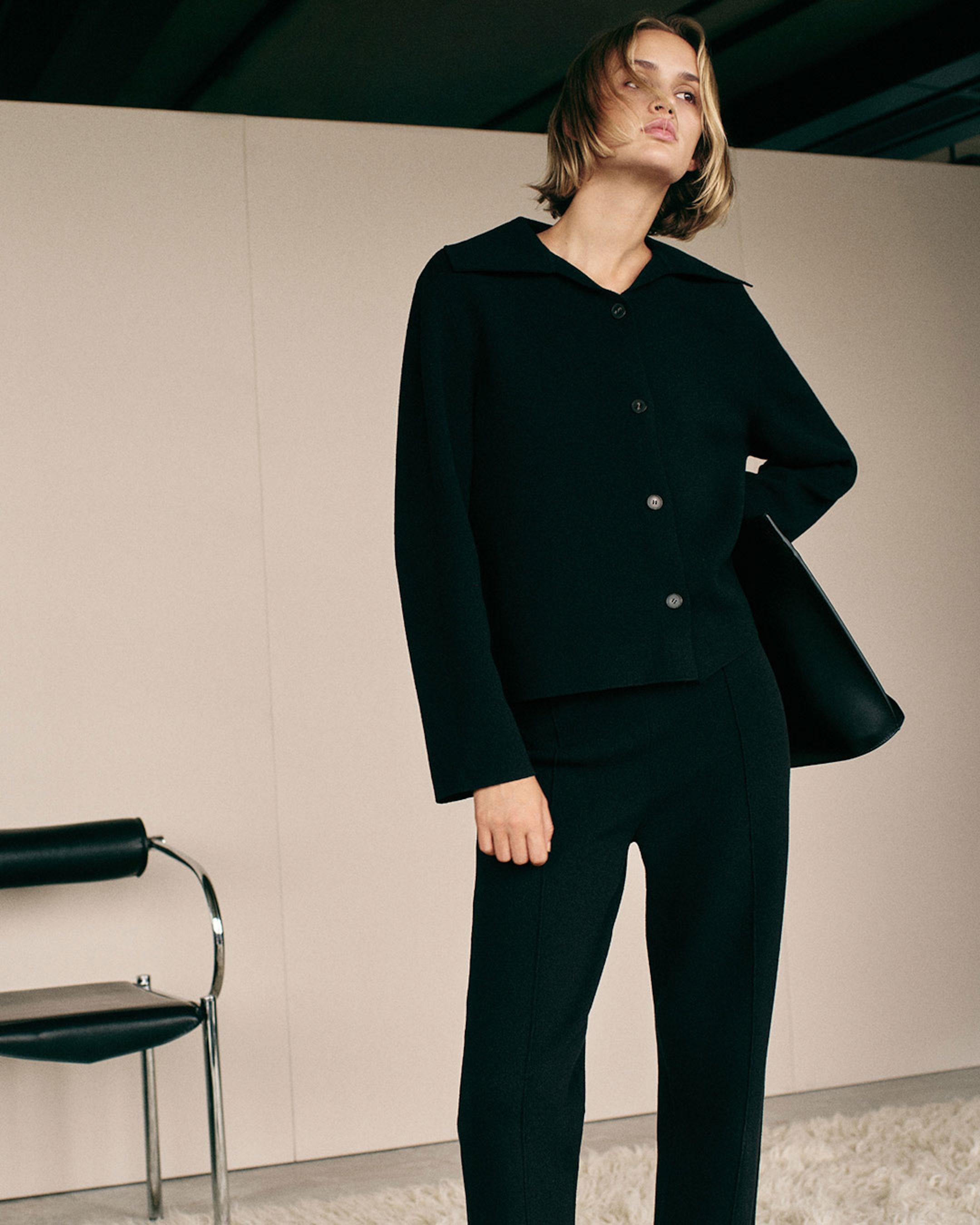 Model in a black tailored outfit stands confidently in a minimalist room with a leather chair and a shaggy white rug.