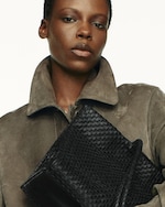 Placeholder for: Close-up of a person in a suede coat holding a black woven leather clutch, neutral expression.
