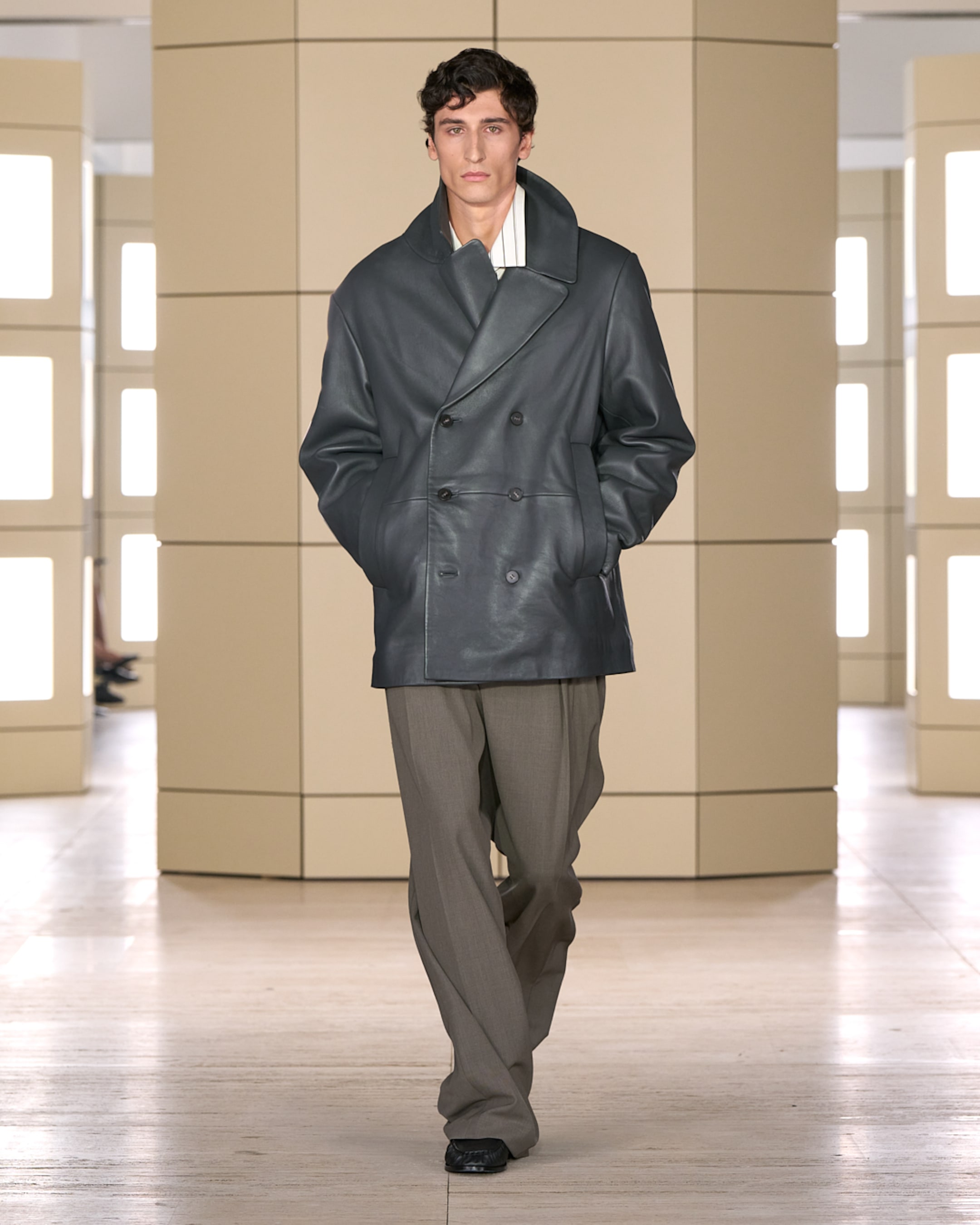 Model wearing a dark, double-breasted coat with hands in pockets, paired with loose brown pants, walking in a minimalist setting.