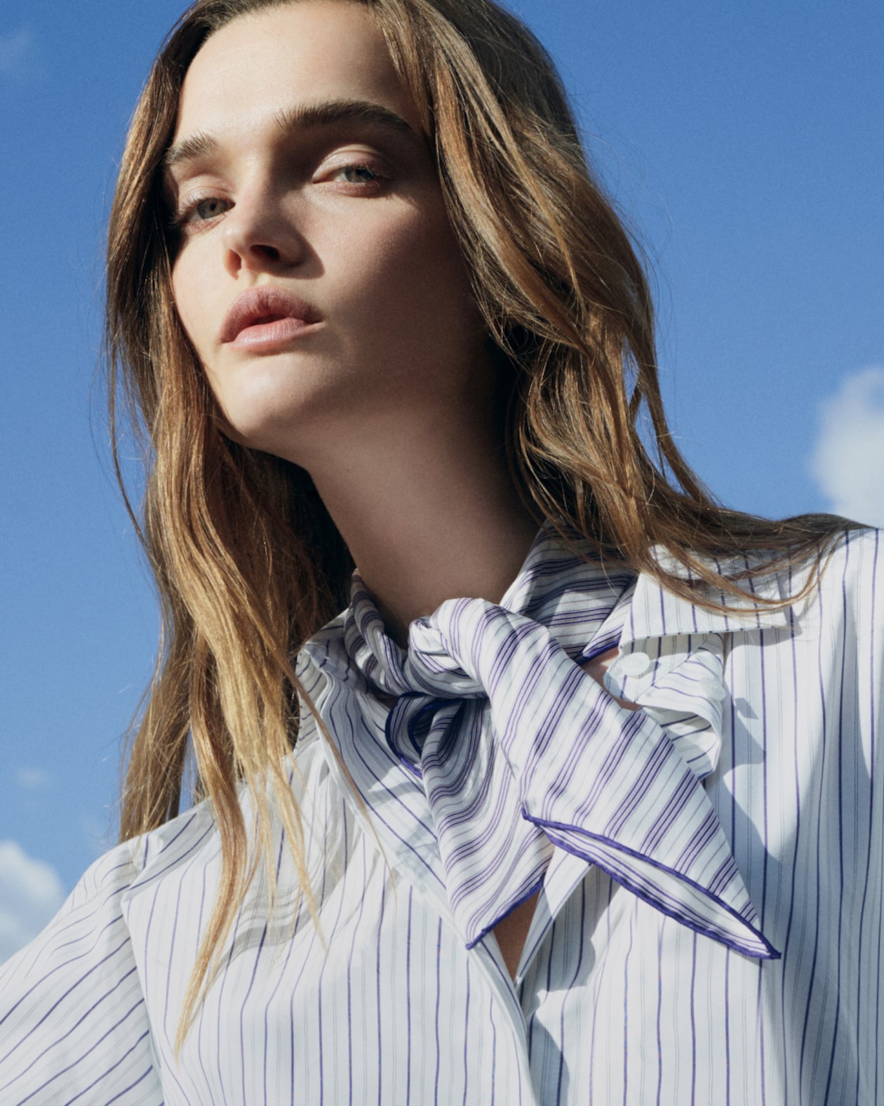 A person with long hair wearing a striped shirt and scarf looks forward under a blue sky with scattered clouds.