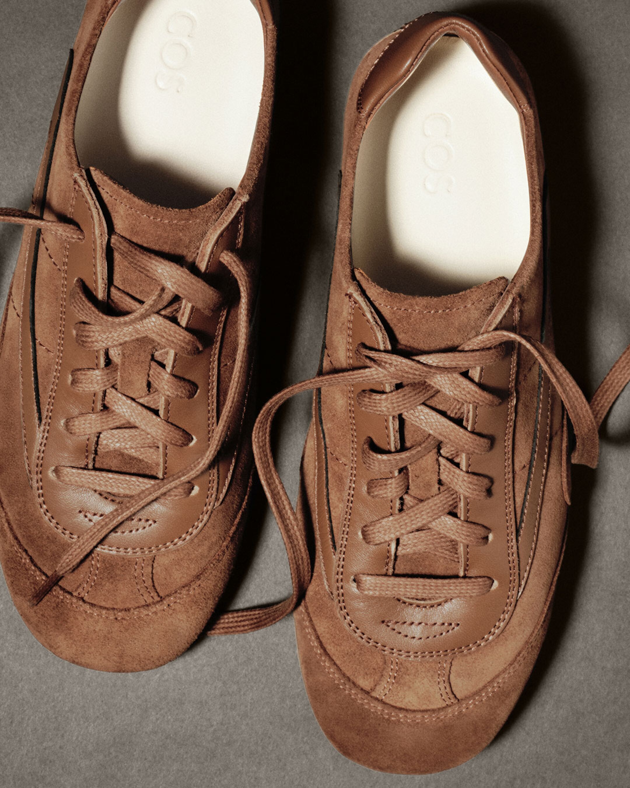 Brown suede sneakers with laces on a gray surface, featuring stitched details and a white inner sole with "COS" branding.