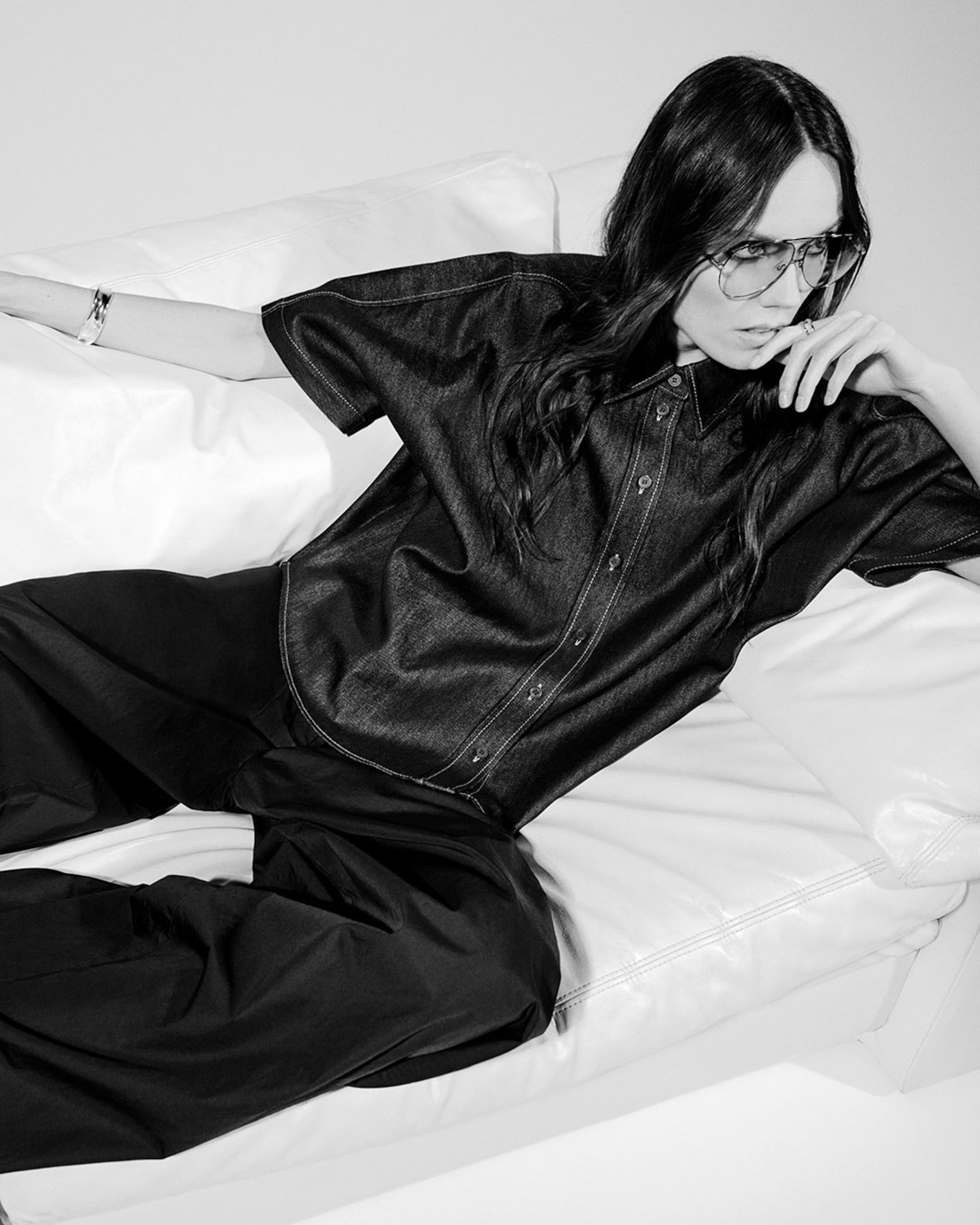 Person in a black button-up shirt and pants, wearing glasses, seated on a white couch with a thoughtful expression. Black and white image.