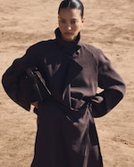 Placeholder for: A person in a dark trench coat stands confidently in a sandy outdoor setting, holding a matching handbag under one arm.