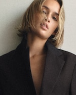 Placeholder for: Close-up portrait of a young woman with short blonde hair in a dark blazer, head tilted, loose strands partially covering her face.
