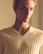 Placeholder for: Close-up portrait of a blond young man in a cream ribbed V-neck sweater, soft warm lighting and neutral background.