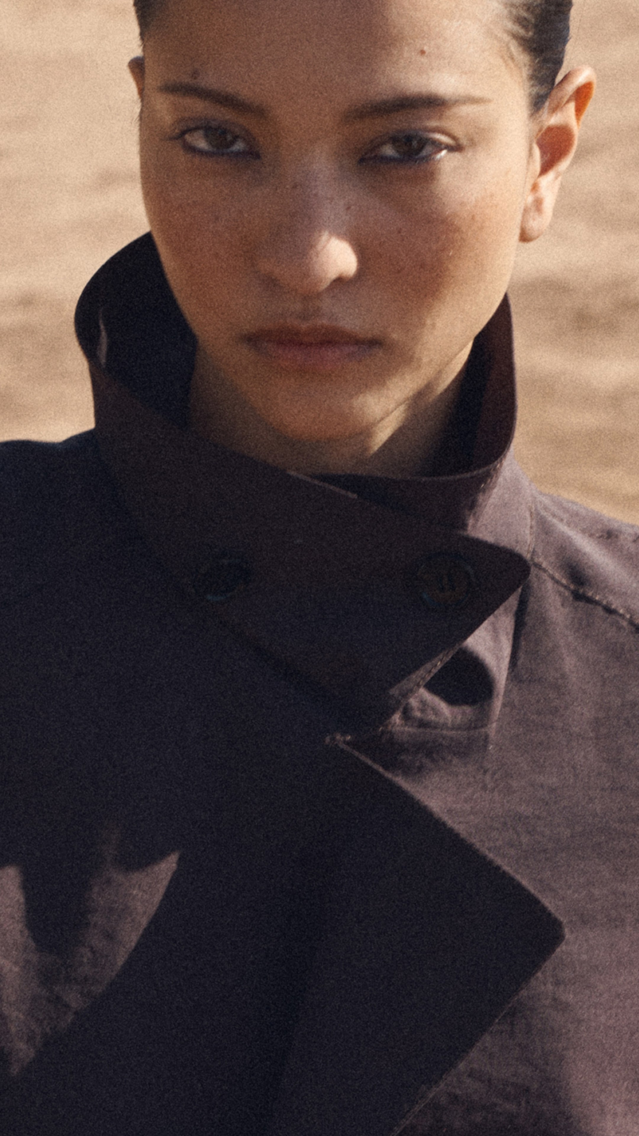 Person in a dark coat with a high collar, looking directly at the camera with a serious expression against a blurred outdoor background.