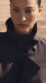 Placeholder for: Person in a dark coat with a high collar, looking directly at the camera with a serious expression against a blurred outdoor background.