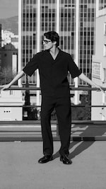 Placeholder for: Black and white photo of a person in sunglasses leaning on a railing on a rooftop, with tall buildings in the background.