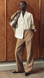 Placeholder for: A person posing confidently in a pinstriped shirt and tan trousers, holding a jacket over their shoulder against a wooden backdrop.