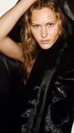 Placeholder for: A person with long wavy hair poses with a confident expression, partially wrapped in a shiny black fabric.