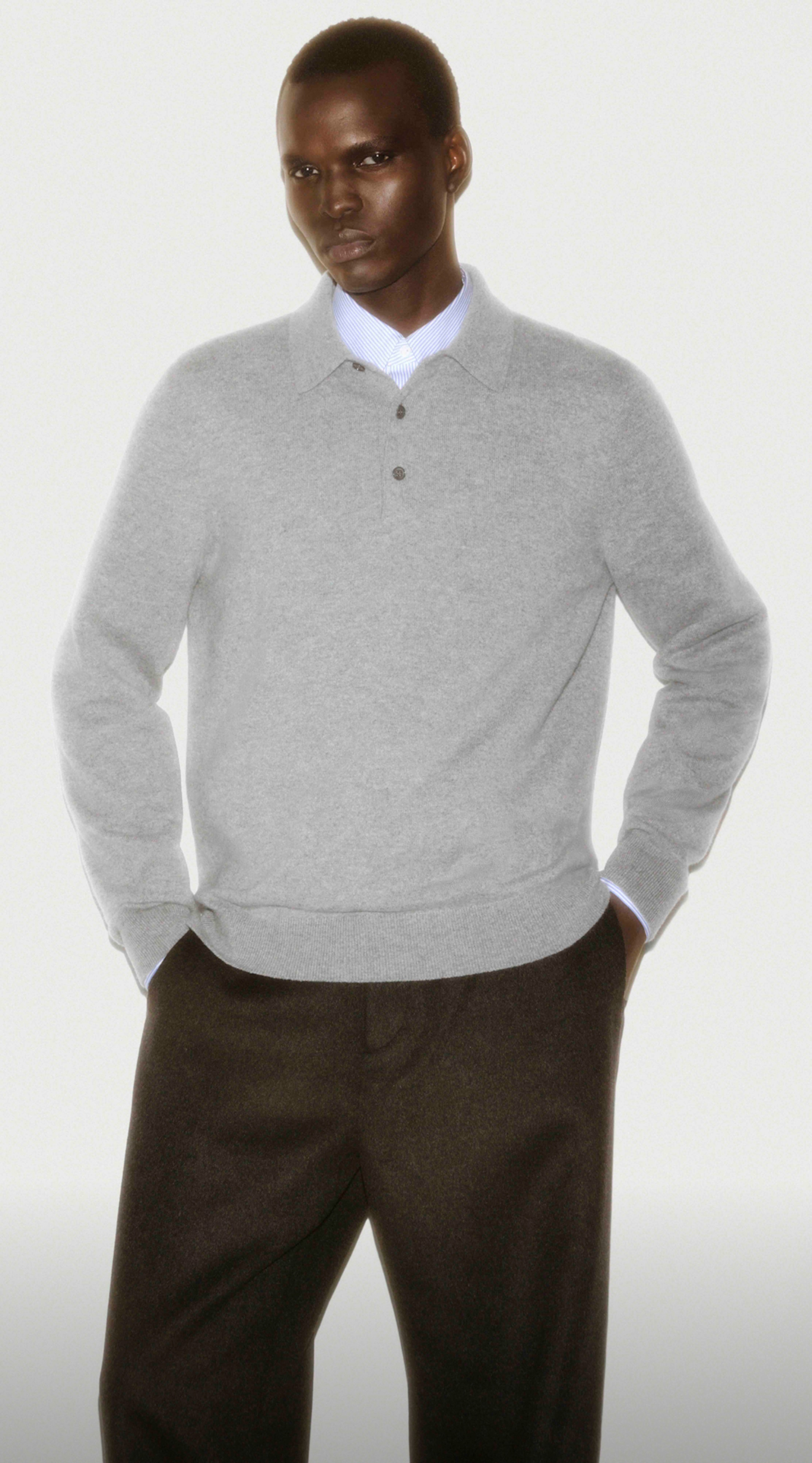 Person wearing a gray sweater over a white shirt and dark trousers, standing with hands in pockets against a plain background.