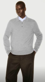 Placeholder for: Person wearing a gray sweater over a white shirt and dark trousers, standing with hands in pockets against a plain background.