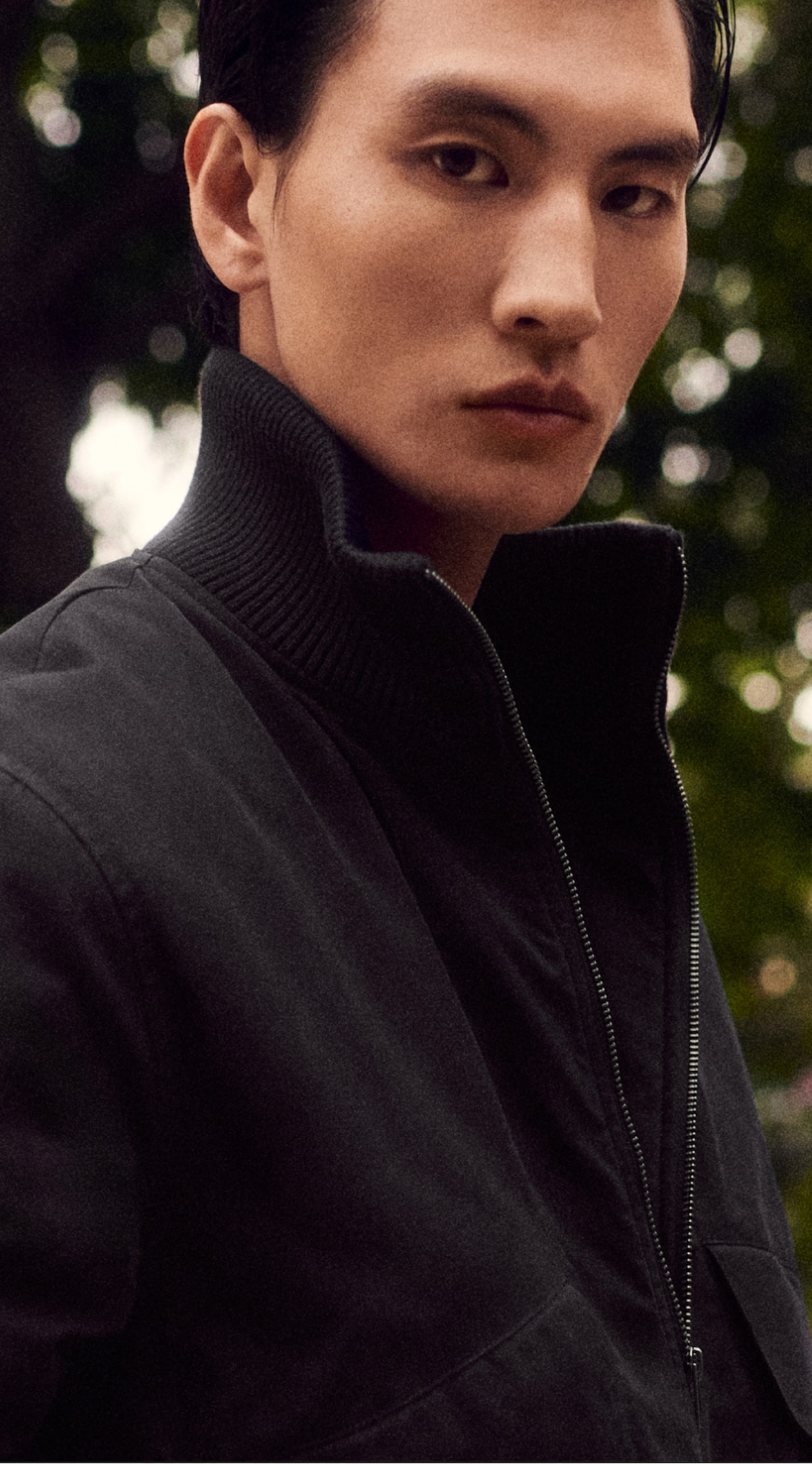 Person in a dark jacket with a high collar, standing outdoors, looking at the camera with a serious expression. Blurred greenery in the background.