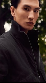 Placeholder for: Person in a dark jacket with a high collar, standing outdoors, looking at the camera with a serious expression. Blurred greenery in the background.