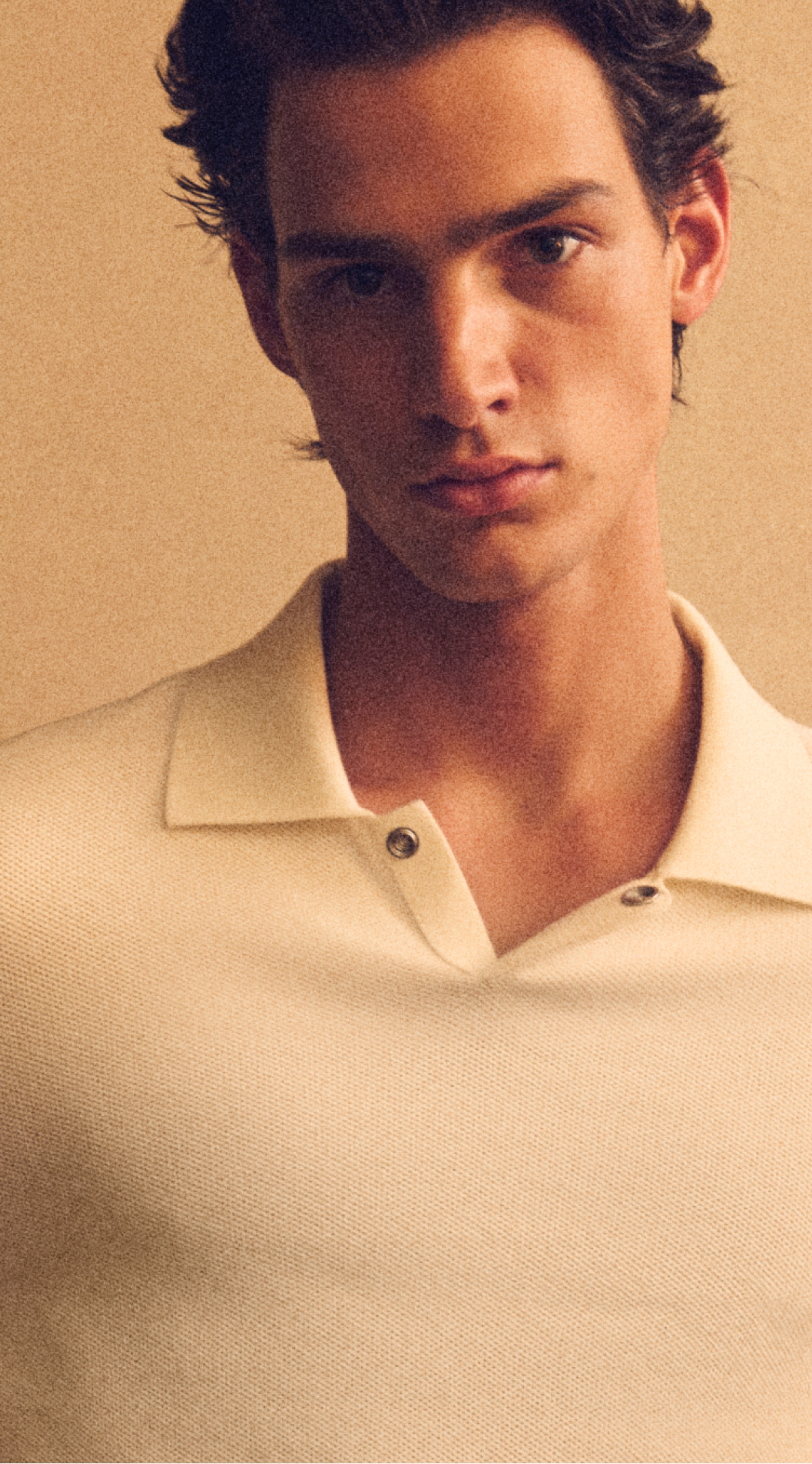 Close-up portrait of a young man with a neutral expression in a cream polo against a warm beige background.