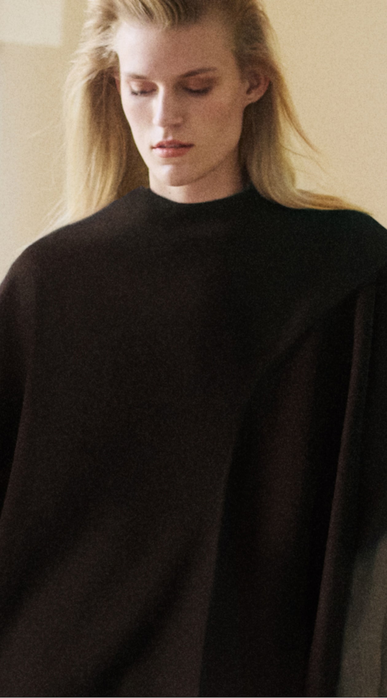 Person with long blonde hair wearing a dark, draped garment, looking down thoughtfully in a softly lit room.