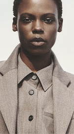 Placeholder for: Close-up portrait of person with short hair, neutral expression, wearing gray button-up shirt and matching blazer against a light background.