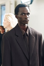 Placeholder for: A man wearing a dark brown suit, shirt, and tie stands confidently indoors. A woman with blonde hair is blurred in the background.
