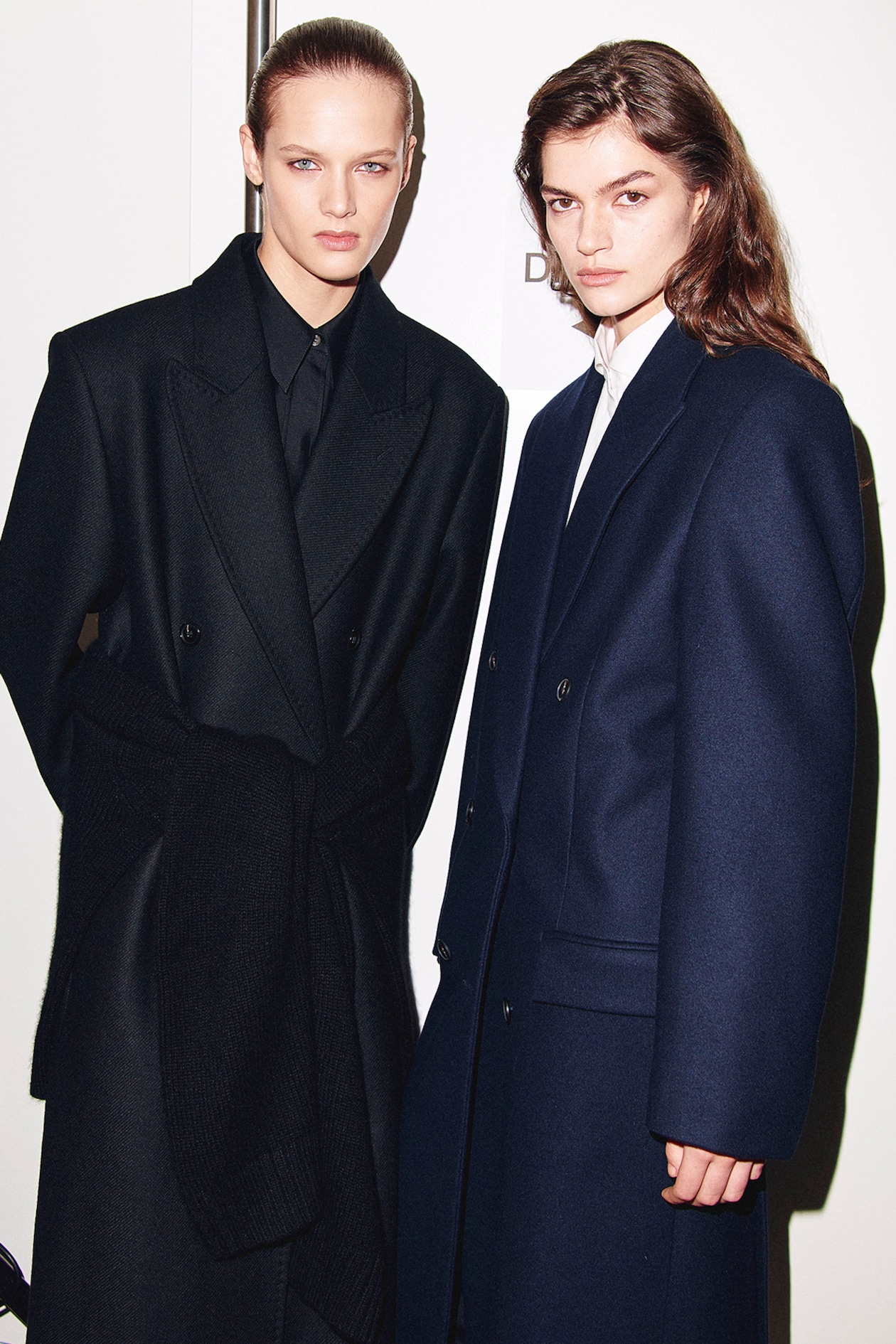 Two individuals in formal dark coats pose against a plain background. They have serious expressions and sleek hairstyles.