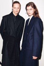 Placeholder for: Two individuals in formal dark coats pose against a plain background. They have serious expressions and sleek hairstyles.
