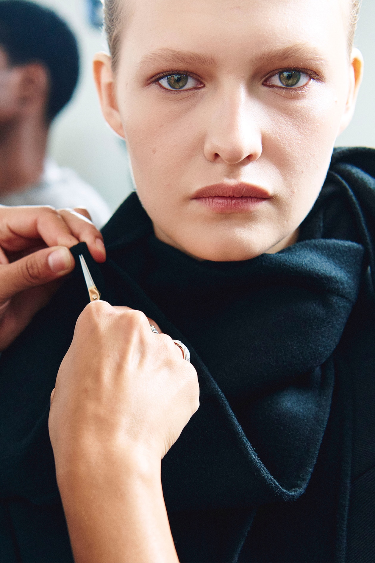 A person staring intently at the camera while another hand adjusts their black clothing with a pin.