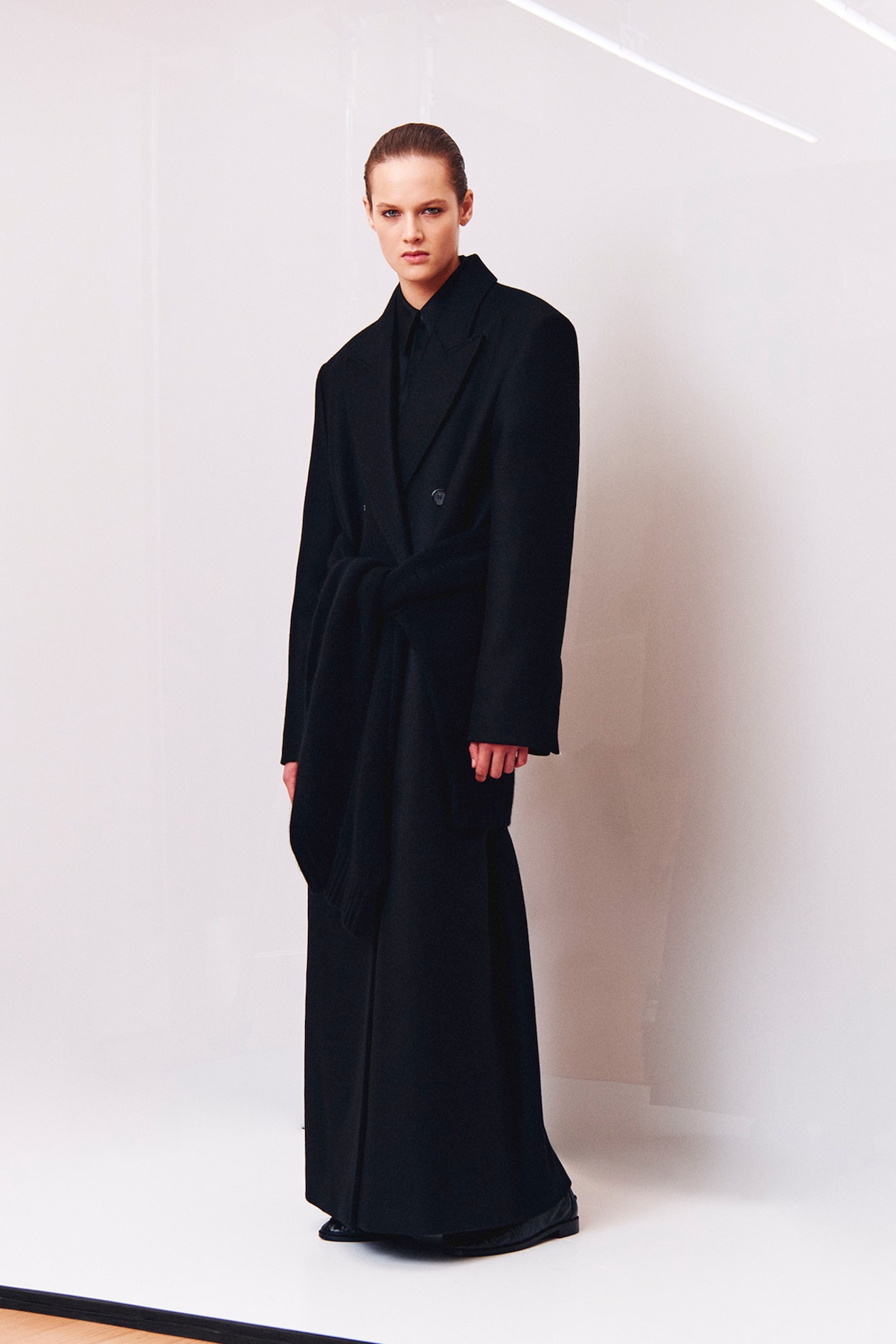 A person stands in a minimalistic setting, wearing an oversized black coat with a tied detail, against a light background.
