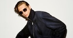 Placeholder for: Man in a dark jacket and sunglasses poses against a white background, with slicked-back hair and a confident expression.