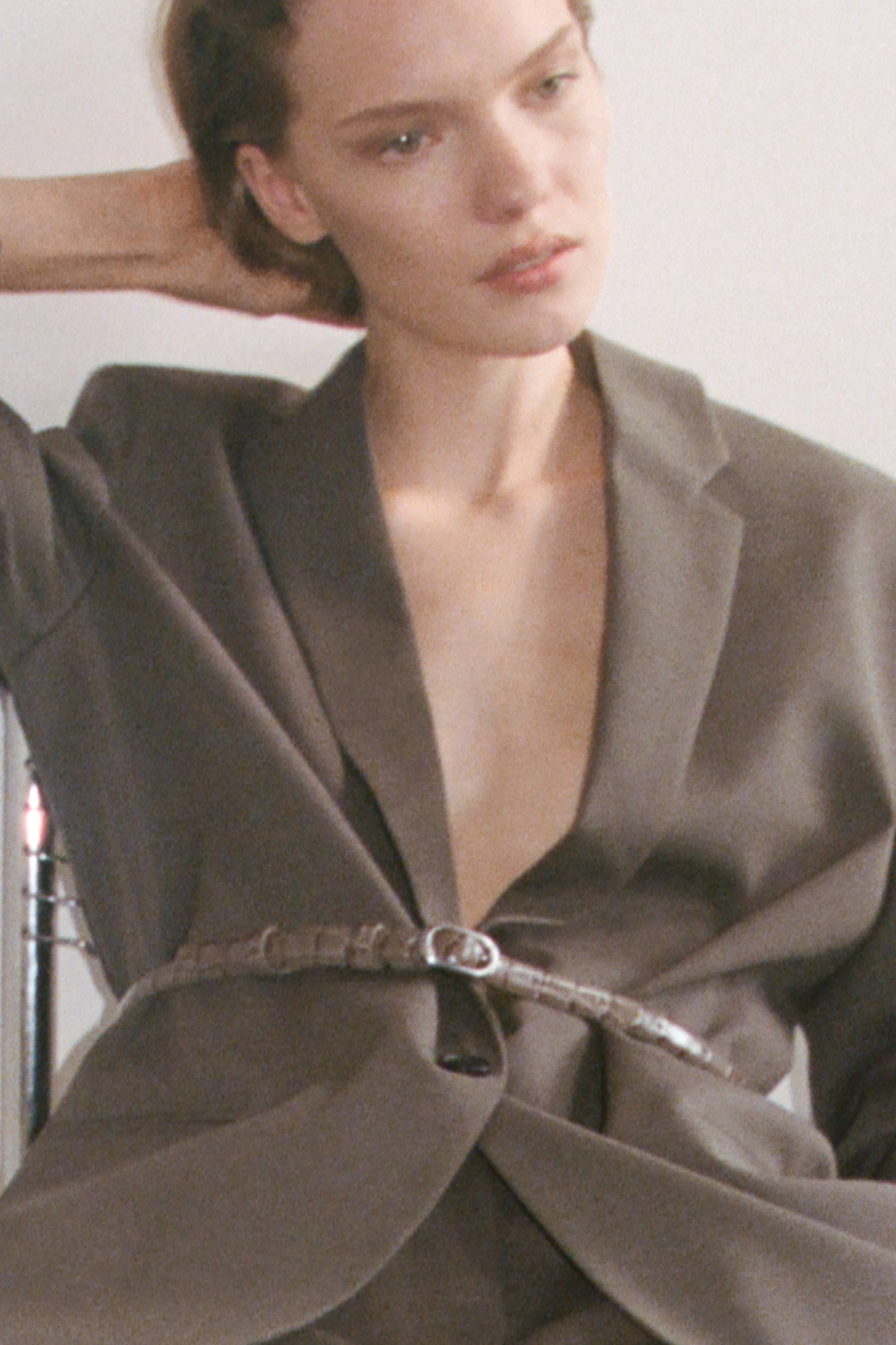 Person in a loose-fitting gray suit with a belt, sitting and looking to the side, with one hand behind their head against a plain background.