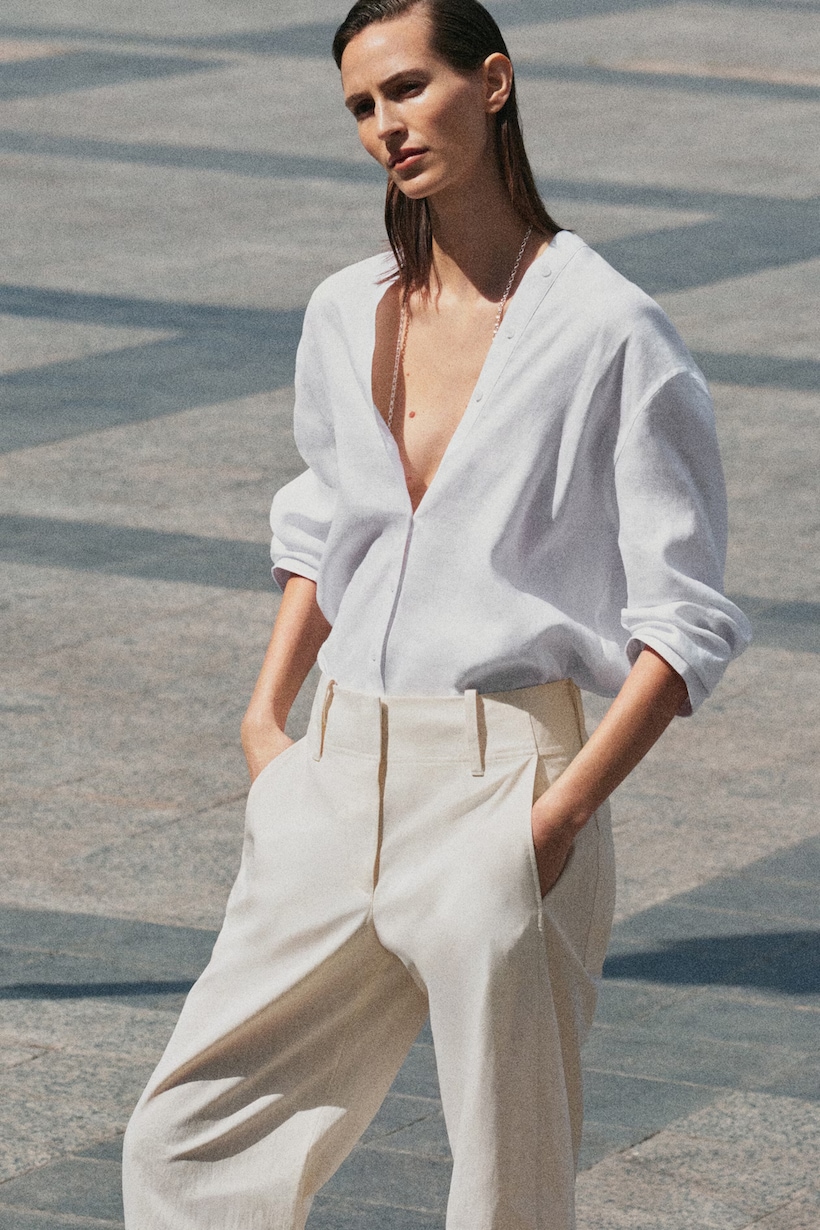 A person wearing a loosely buttoned white shirt and beige trousers, standing outdoors on a stone-paved surface.