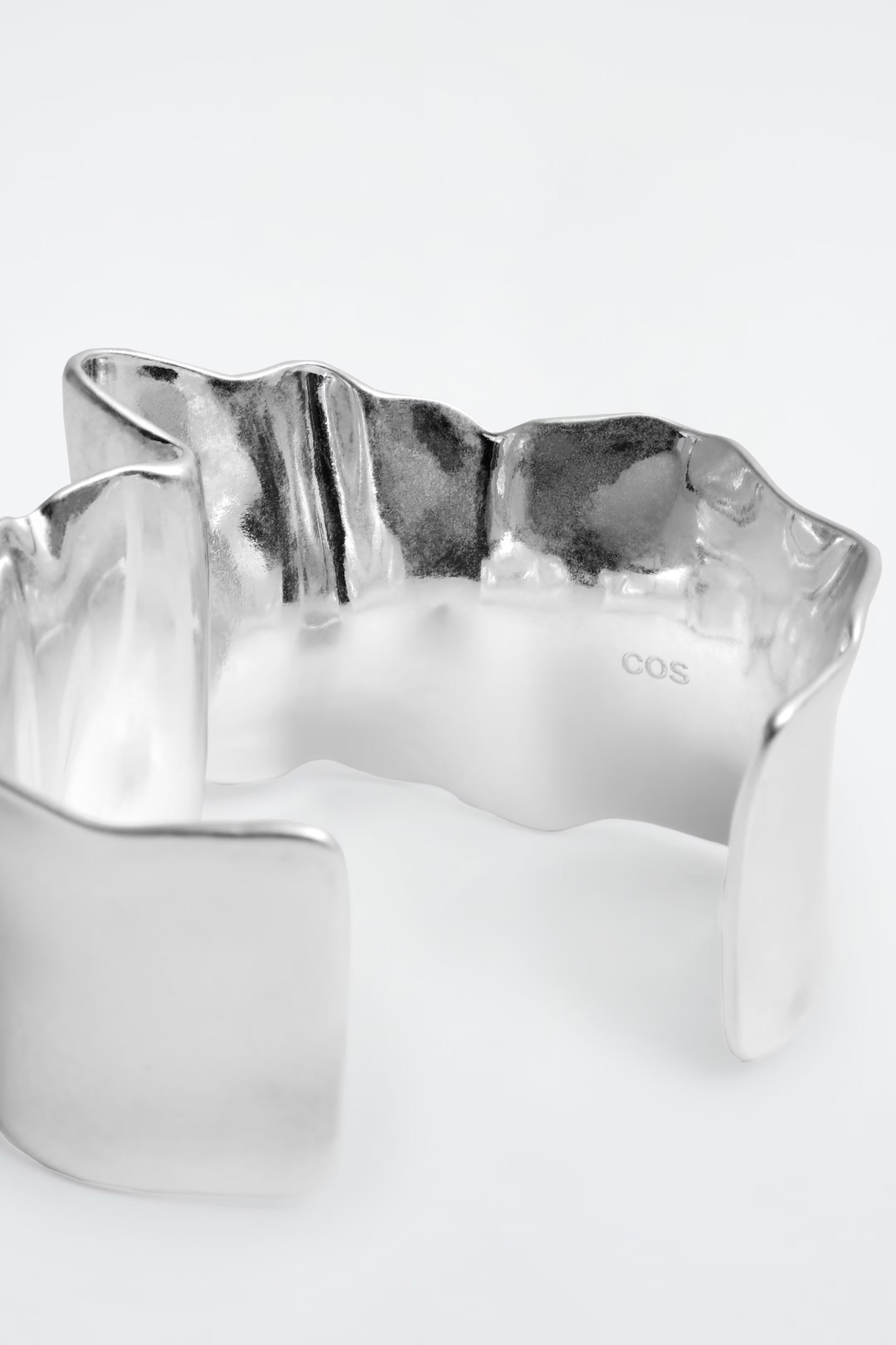 A wavy, metallic silver cuff bracelet with a smooth, reflective surface and subtle engraving on the inside.