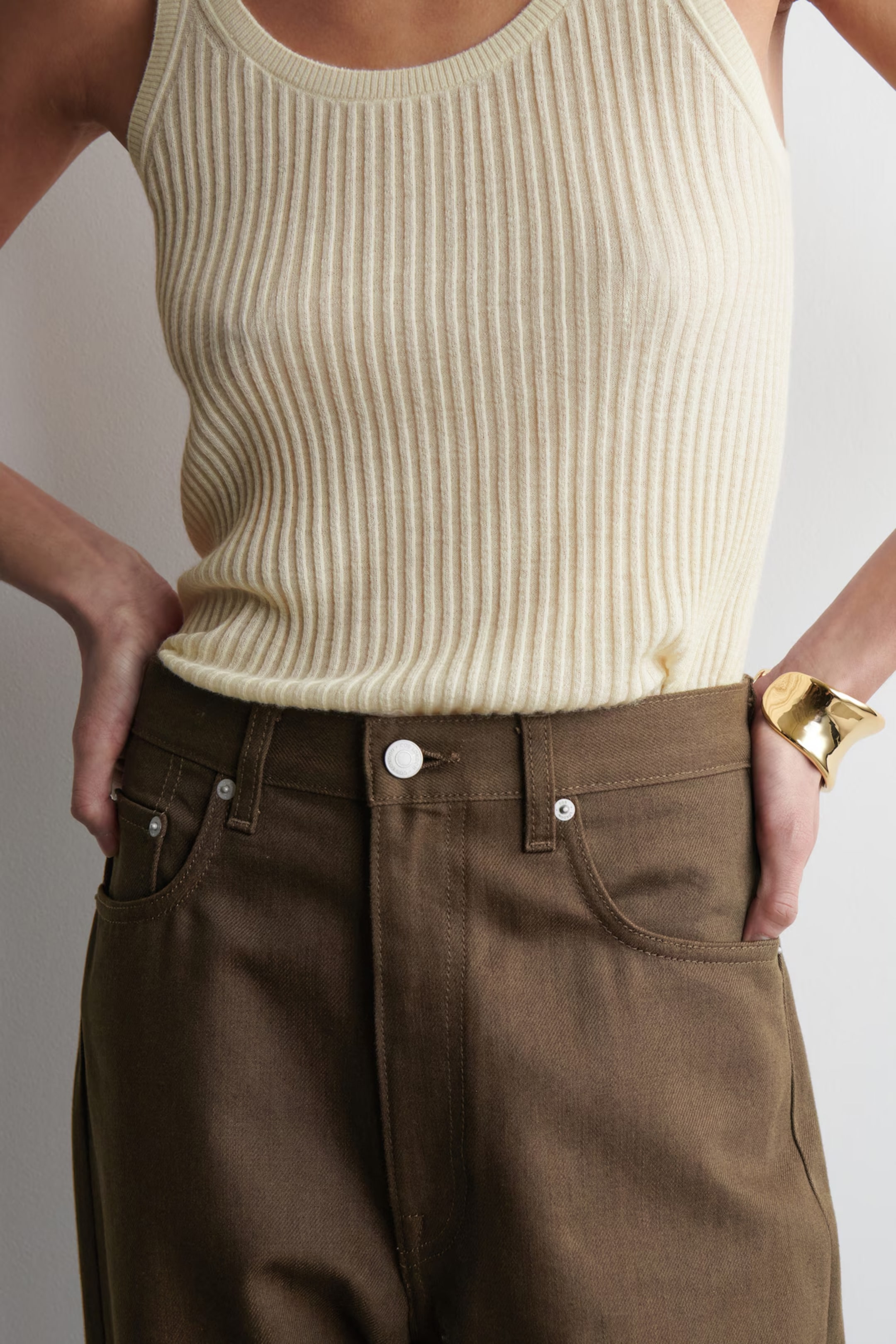 Person wearing a ribbed beige tank top, brown pants, and a gold bracelet, standing with hands on hips against a plain background.