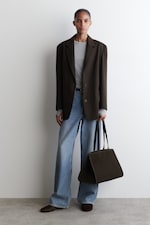 Placeholder for: Person wearing a dark blazer, gray shirt, and wide-leg jeans, holding a large dark tote bag, stands against a plain white background.