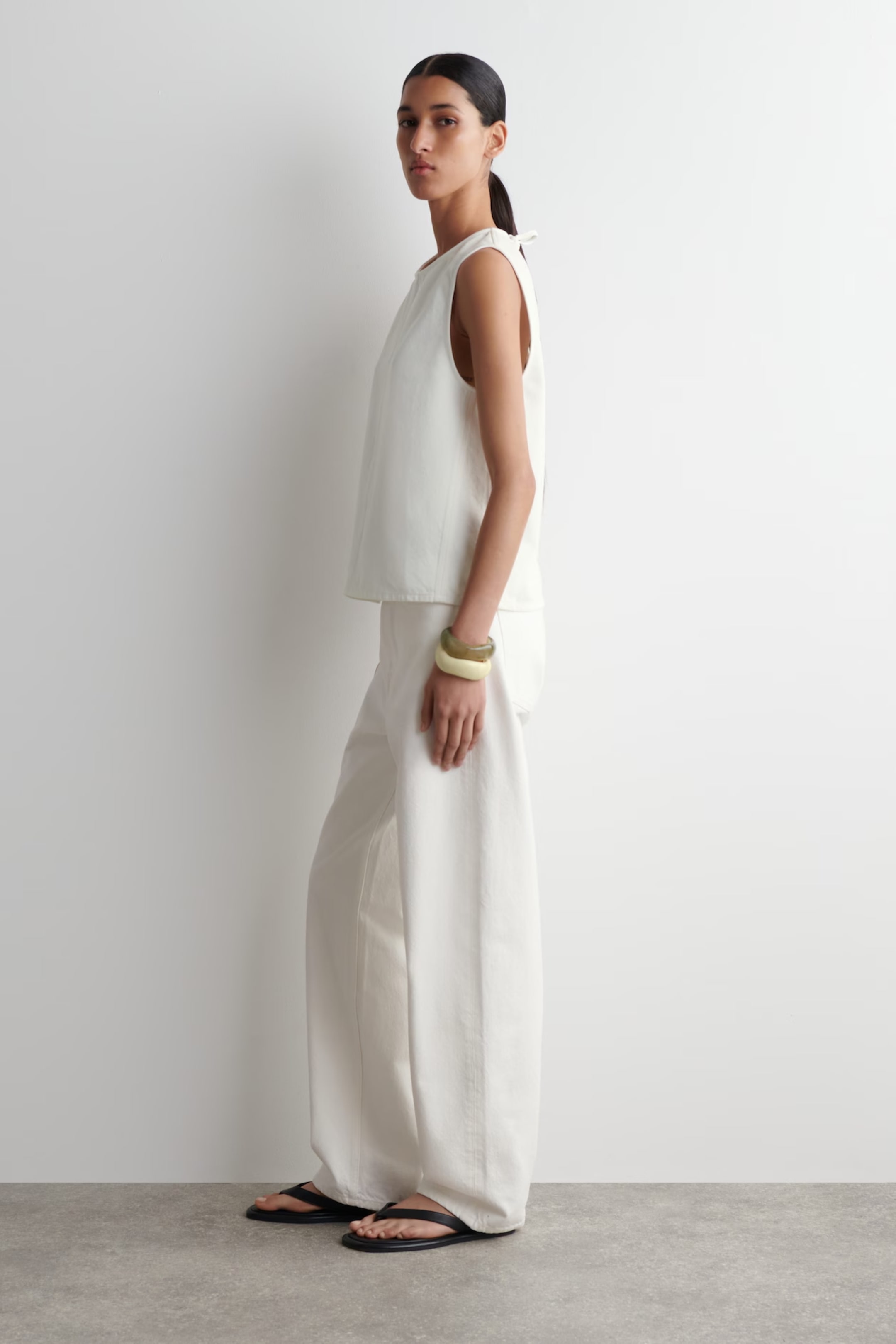 Woman in white sleeveless top and wide-leg pants, wearing black sandals and bracelets, standing sideways against a plain white background.