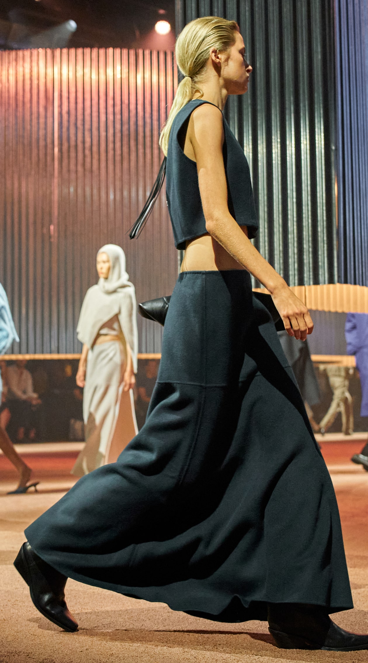 Model walking on a runway wearing a dark, sleeveless top and long skirt. Others in similar attire are in the background.